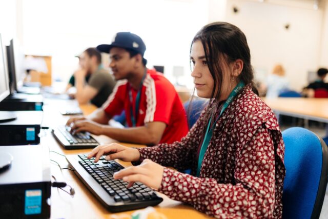 Students focused at computer workstations in a classroom. A young woman in a patterned shirt types, with others nearby. The mood is studious.