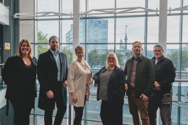 A group of six people stand smiling in front of a large window showcasing an urban view with buildings and trees, conveying a professional and friendly atmosphere.