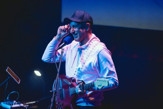 A musician in a hoodie and cap plays an electric guitar on stage, smiling, under dynamic lighting, creating a relaxed and engaging atmosphere.