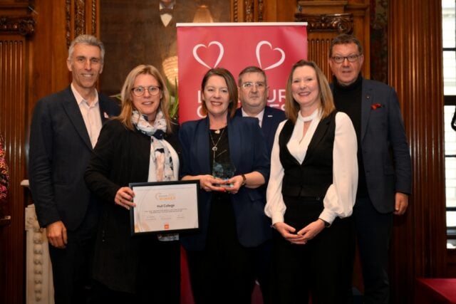 A group of six people smiles while posing with an award. In the background, a red banner with hearts adds a celebratory atmosphere. They appear happy and proud.