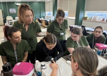 A group of women in green uniforms attentively watch a nail technician demonstrating a manicure technique. The setting is a bright classroom.