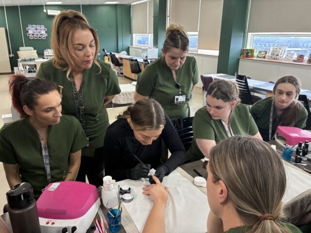 A group of women in green uniforms attentively watch a nail technician demonstrating a manicure technique. The setting is a bright classroom.