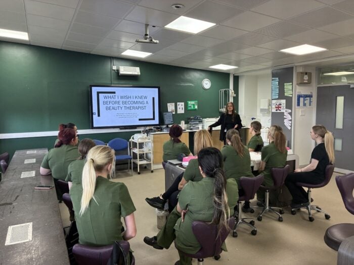 Classroom with students in green uniforms facing a teacher presenting “What I Wish I Knew Before Becoming a Beauty Therapist” on a screen. Engaged atmosphere.