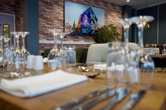 Restaurant interior with glassware on a wooden table. A large mural depicts urban art on a brick wall, adding a vibrant, modern touch. Warm lighting and casual ambiance.