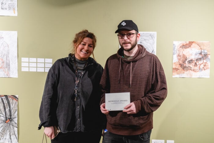 Two people stand smiling in front of a green wall displaying artwork. The person on the right holds a plaque reading "Monocle Award," suggesting an art event.