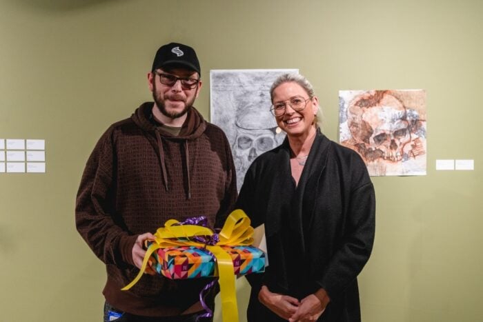 A man in a brown hoodie and cap holds a colourful gift with bright ribbons, standing next to a smiling woman in black. Two skull artworks on the wall behind them.