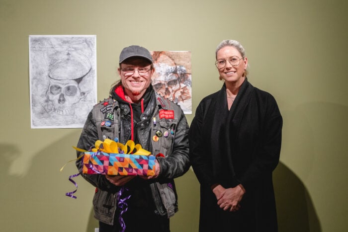 Two people stand smiling in front of two artworks on a green wall. The person on the left holds a colourful wrapped gift, wearing a jacket with patches.