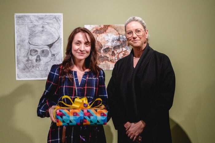 Two women stand smiling in front of skull drawings on a light green wall. One holds a colorful gift with a yellow ribbon, creating a cheerful atmosphere.