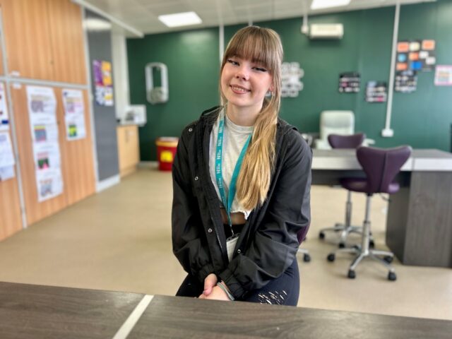 Smiling young woman with long hair, wearing a black jacket and teal lanyard, sits in a classroom with green walls, posters, and office chairs.