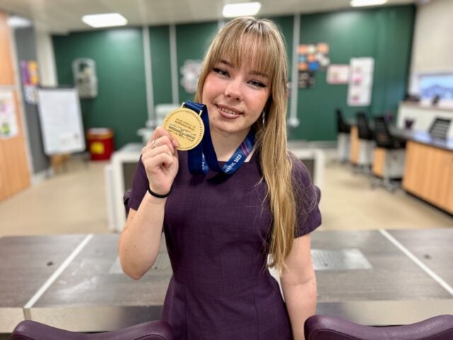 A woman in a purple dress smiles, holding a gold medal with a blue ribbon, in a classroom with wooden desks and a green wall. Celebratory mood.