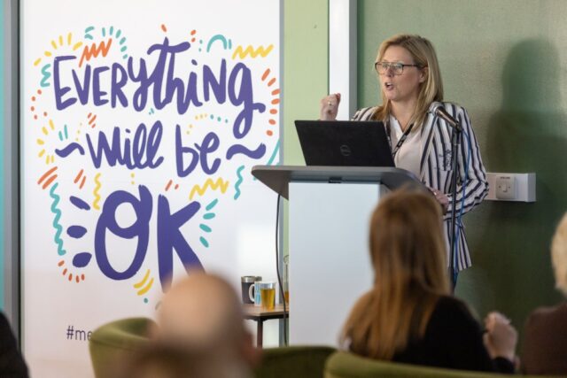 A woman speaks at a podium with a laptop in front of a sign saying "Everything will be OK" in colorful letters. Audience members in foreground.