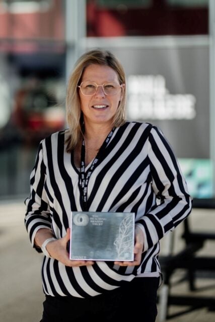 A smiling woman in glasses and a black-and-white striped shirt holds an award plaque. The background shows a blurred modern building.