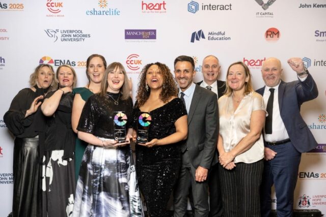 A group of smiling people poses on a red carpet, two women holding awards. The backdrop displays various sponsor logos. The mood is celebratory.