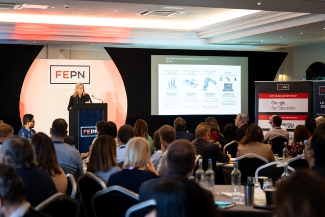A presenter speaks at a conference, standing at a podium with a large screen displaying a presentation. Attentive audience members fill the room.