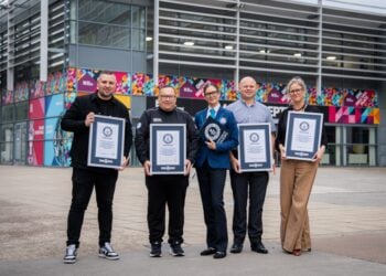 Five people stand smiling in front of a modern building with colorful art, each holding a framed certificate, suggesting a celebratory or award event.