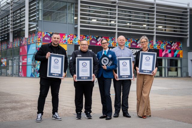 Five people stand smiling in front of a modern building with colorful art, each holding a framed certificate, suggesting a celebratory or award event.