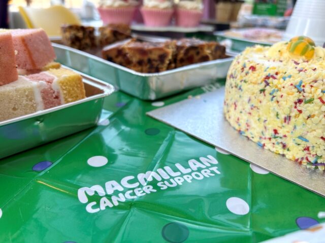 Cakes on a table with green Macmillan Cancer Support cloth, including a sprinkle-covered cake, Battenberg, and cupcakes, creating a cheerful, supportive vibe.