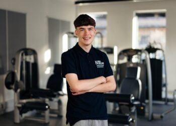 A male Sport student at Hull College stands smiling in a gym, arms crossed. Fitness equipment is visible in the bright, modern space behind him.