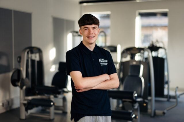 A male Sport student at Hull College stands smiling in a gym, arms crossed. Fitness equipment is visible in the bright, modern space behind him.