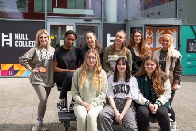 A group of nine young adults, smiling outside Hull College. They are casually dressed, standing and seated in front of a modern building, conveying a cheerful and friendly atmosphere.