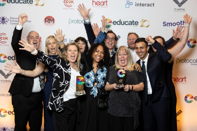 A group of joyful people, dressed in formal attire, celebrates with hands raised and smiles, holding colourful awards against a backdrop of sponsor logos.