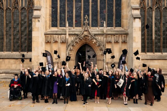 Graduates in black robes cheerfully toss caps in the air outside a historic stone building, celebrating their achievement. The mood is joyful and triumphant.