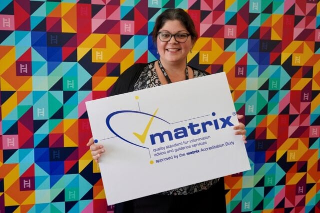 Smiling person holds a "matrix" accreditation sign in front of a colourful geometric background, conveying professionalism and positivity.