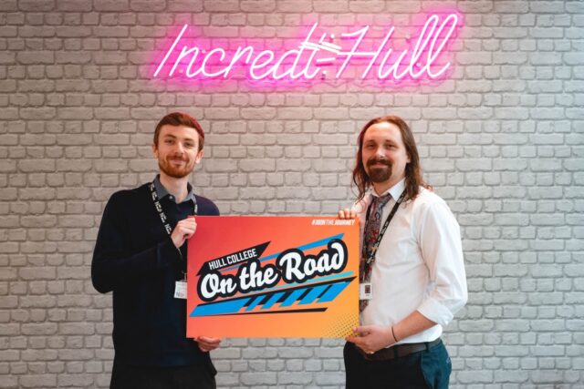 Two men stand smiling, holding a colourful sign reading "Hull College On the Road." A neon "Incredi-Hull" sign glows on the brick wall behind them.
