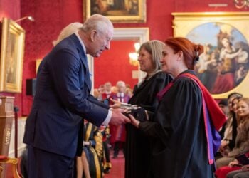 A distinguished man in a blue suit shakes hands with a woman in academic robes inside an ornate room with red walls and classic paintings.