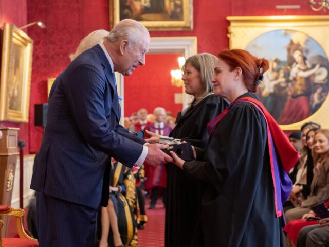 A distinguished man in a blue suit shakes hands with a woman in academic robes inside an ornate room with red walls and classic paintings.