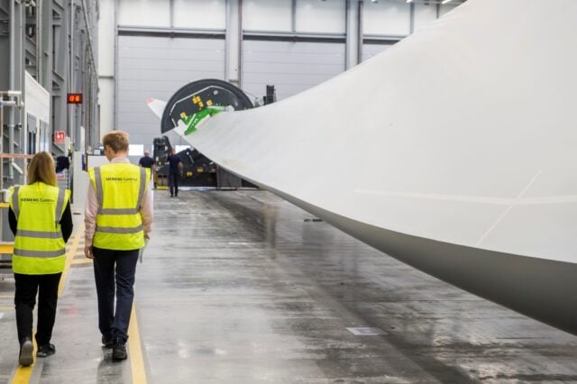 Two workers wearing high-visibility vests walk beside a massive wind turbine blade inside an expansive, clean factory setting, conveying innovation and scale.