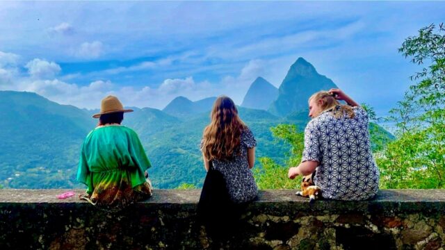 Three people sit on a stone wall, overlooking lush green mountains with a blue sky and scattered clouds. The scene conveys tranquility and wonder.