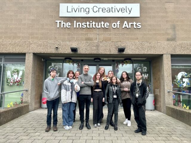A group of ten people stands smiling in front of the Institute of Arts building. The mood is cheerful and welcoming, with modern architecture in the background.