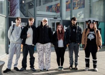 Six individuals stand in a line outside a modern building. They are wearing casual and varied clothing, exuding a relaxed and confident vibe.