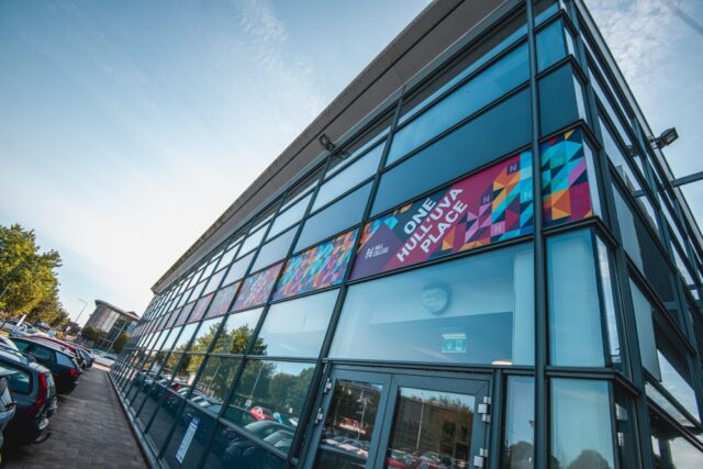 Modern glass building exterior with colorful geometric signage reading "One Hull'uva Place." Cars are parked alongside a tree-lined street under a clear blue sky.