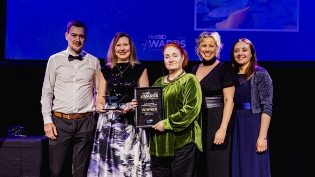 A group of five people stands on a stage at an awards ceremony. They are smiling and holding a winner's plaque. The background is dark with blue lighting.