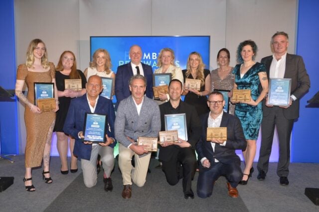A group of 13 people, dressed formally, pose proudly with plaques and awards on a stage. The background shows a banner reading "Plumbing & Heating New Installer Awards," creating a celebratory atmosphere.