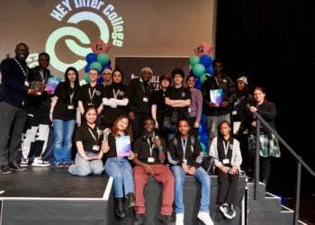 A diverse group of young people in black shirts poses on stage at HEY Inter College. They hold colourful certificates, smiling amid a celebratory atmosphere.