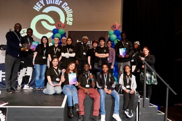 A diverse group of young people in black shirts poses on stage at HEY Inter College. They hold colourful certificates, smiling amid a celebratory atmosphere.