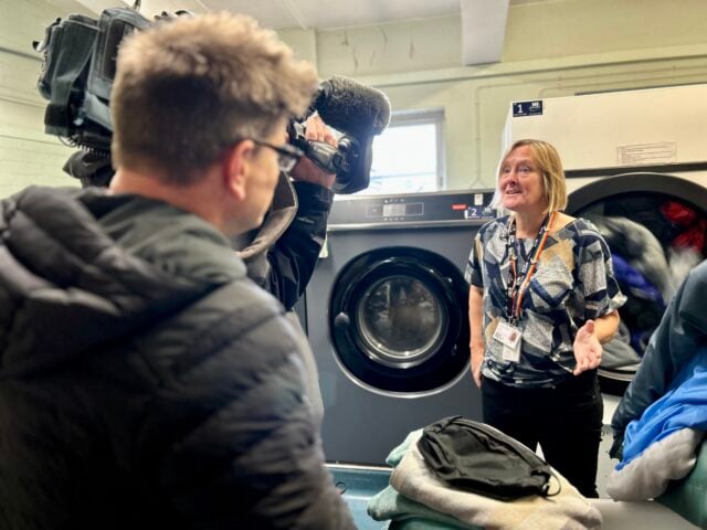 A woman in a patterned shirt speaks to a cameraman in a laundry setting. She appears animated and engaged, with large machines in the background.