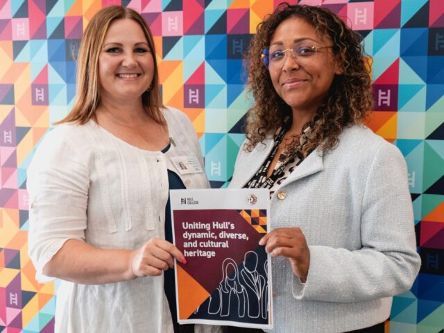 Two women smiling and holding a flyer titled "Uniting Hull's dynamic, diverse, and cultural heritage" against a colourful geometric backdrop.