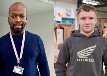 Two men standing indoors, one wearing a blue sweater with an ID lanyard and the other in a grey Honda hoodie. The setting is warm and engaging.