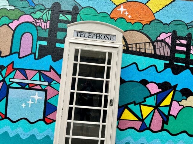 A white telephone booth is set against a vibrant mural depicting a colourful geometric landscape with bridges, waves, and a sun, creating a whimsical tone.