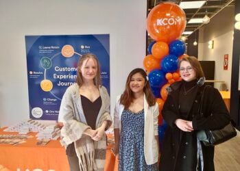 Three women stand smiling at a event held at KCOM. Behind them, a sign and balloon arch in blue and orange. The atmosphere is welcoming.