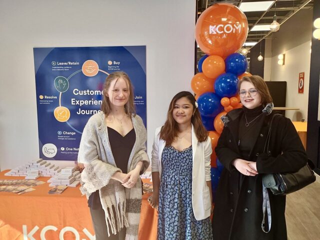Three women stand smiling at a event held at KCOM. Behind them, a sign and balloon arch in blue and orange. The atmosphere is welcoming.