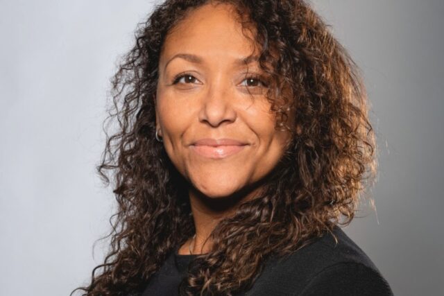 Smiling woman with curly hair in a black top against a neutral gray background. Her expression conveys confidence and warmth.
