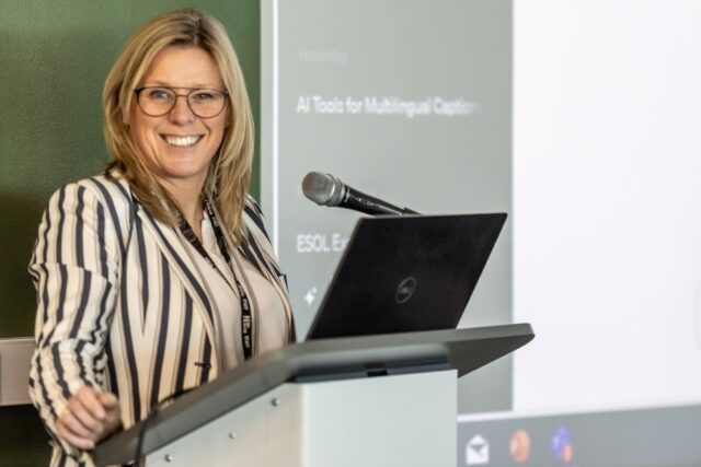 A smiling woman in glasses and a striped blazer stands at a podium with a laptop and microphone, presenting on AI tools for multilingual captioning.