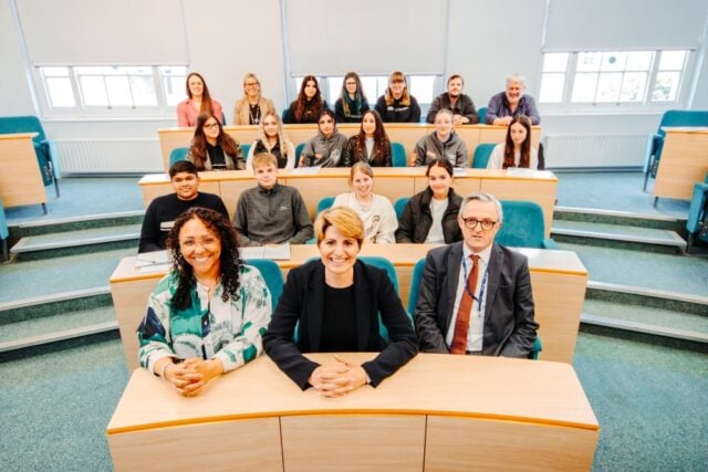 A group photo in a classroom setting shows three professionals seated at the front and students behind. The atmosphere is formal yet friendly.