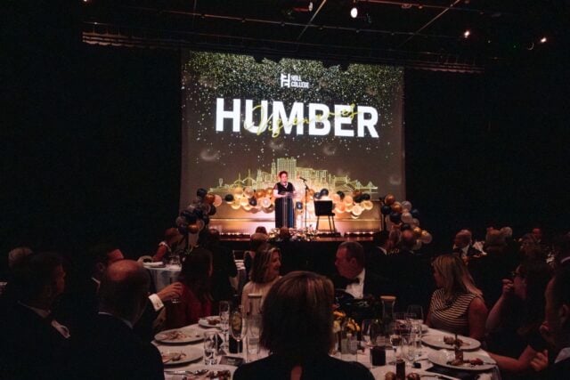 A formal event with a speaker on stage before an audience. The backdrop displays "Humber" with festive lights and gold and blue balloon decorations.