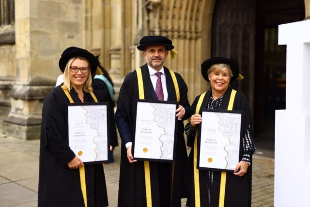 Three people in academic robes and hats hold framed "Order of Distinction" certificates outside a historic building, smiling proudly.